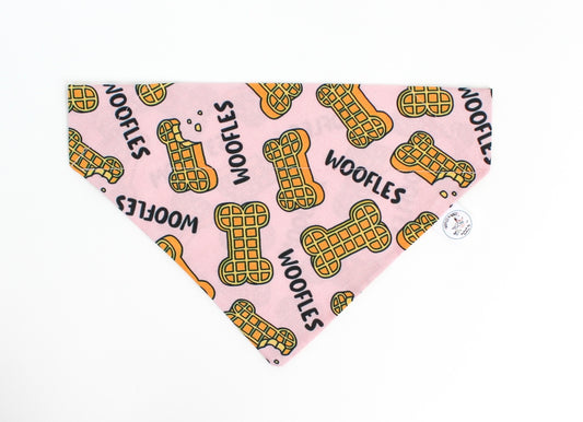 A pink dog bandana featuring a repeating pattern of yellow waffle-shaped bones with black outlines and the word 'WOOFLES' printed in black, shown on a plain white background.