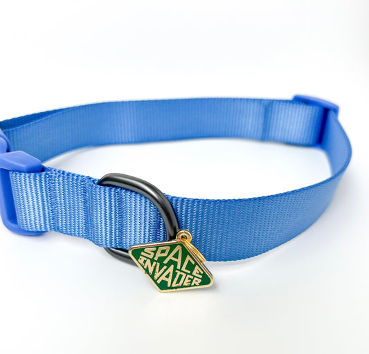 A blue nylon pet collar with a black metal ring and a green diamond-shaped enamel charm that reads 'SPACE INVADER' in gold lettering, shown on a white background.