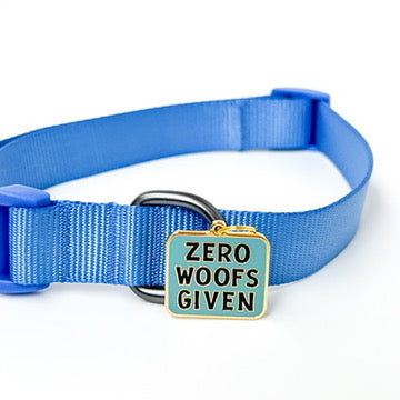 A blue nylon pet collar with a metal ring and a square enamel tag that reads 'ZERO WOOF'S GIVEN' in black text on a light blue background with gold trim, shown on a white background.