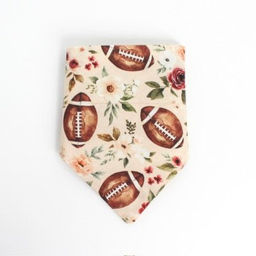 An over the collar bandana featuring a pattern of brown American footballs and watercolor-style flowers and leaves on a light peach background, displayed flat against a white background.