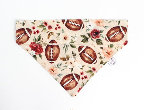 An over the collar pet bandana with a light peach background, featuring a repeating pattern of brown footballs and red and pink roses with green leaves. A small white circular tag is attached to the right side.