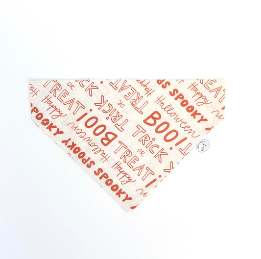 A triangular bandana with a Halloween theme, featuring a cream background with orange text patterns like 'BOO!', 'TRICK or TREAT', 'Happy Halloween', and 'SPooktacular'. A small logo tag is attached near one corner.