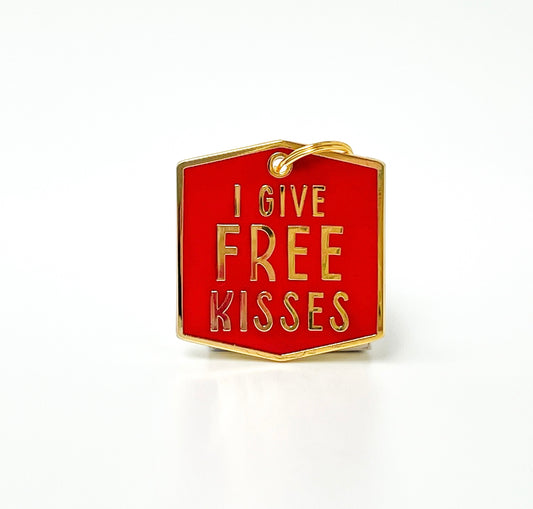 A small hexagonal enamel charm with a red background and gold-tone border and lettering that reads 'I GIVE FREE KISSES', shown on a clean white background.