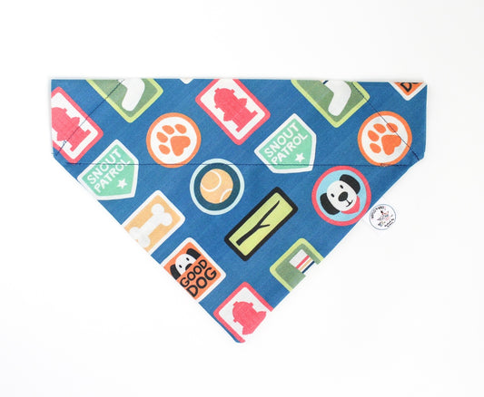 A blue pet bandana featuring a colorful pattern of dog-themed badges, including 'Snout Patrol' and 'Good Dog' designs, laid flat on a white background.