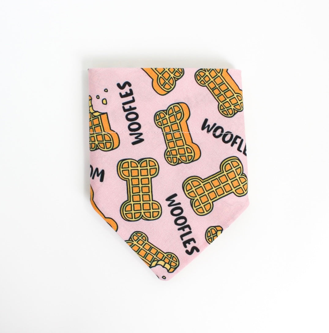 A light pink pet bandana featuring a repeating pattern of yellow waffle-shaped dog bones with black outlines and the word 'WOOFLES' printed in black.