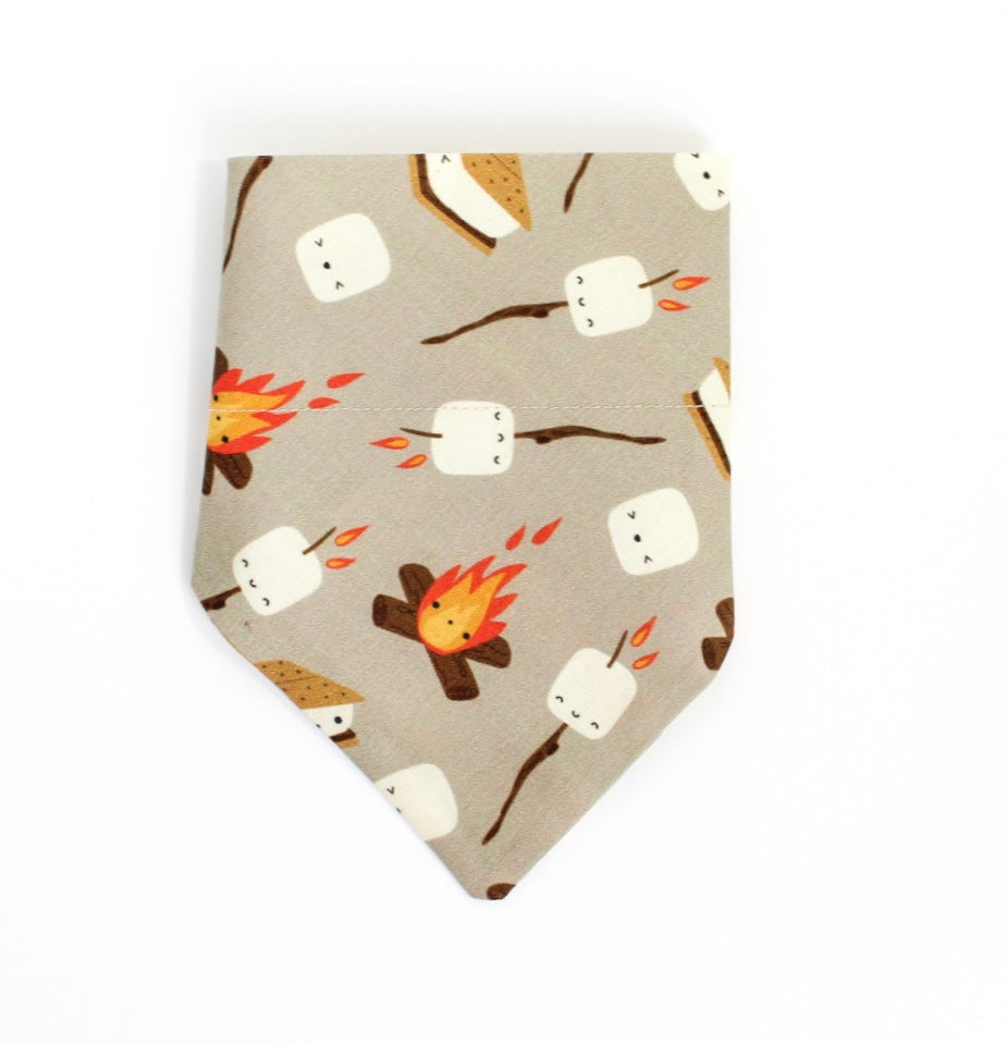 A pet bandana with a s'mores pattern featuring marshmallows, campfires, and graham crackers on a light gray background, shown against a plain white studio backdrop.