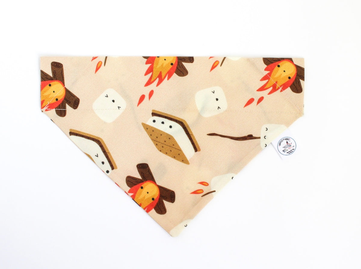 An over the collar pet bandana with a light peach background, featuring a whimsical print of campfires, marshmallows, and s'mores. A small white circular brand tag is attached to the right side, photographed on a clean white background