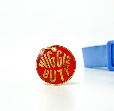 A red oval enamel pin with gold lettering that reads 'WIGGLE BUTT' on a white background, with a blue strap partially visible on the right.
